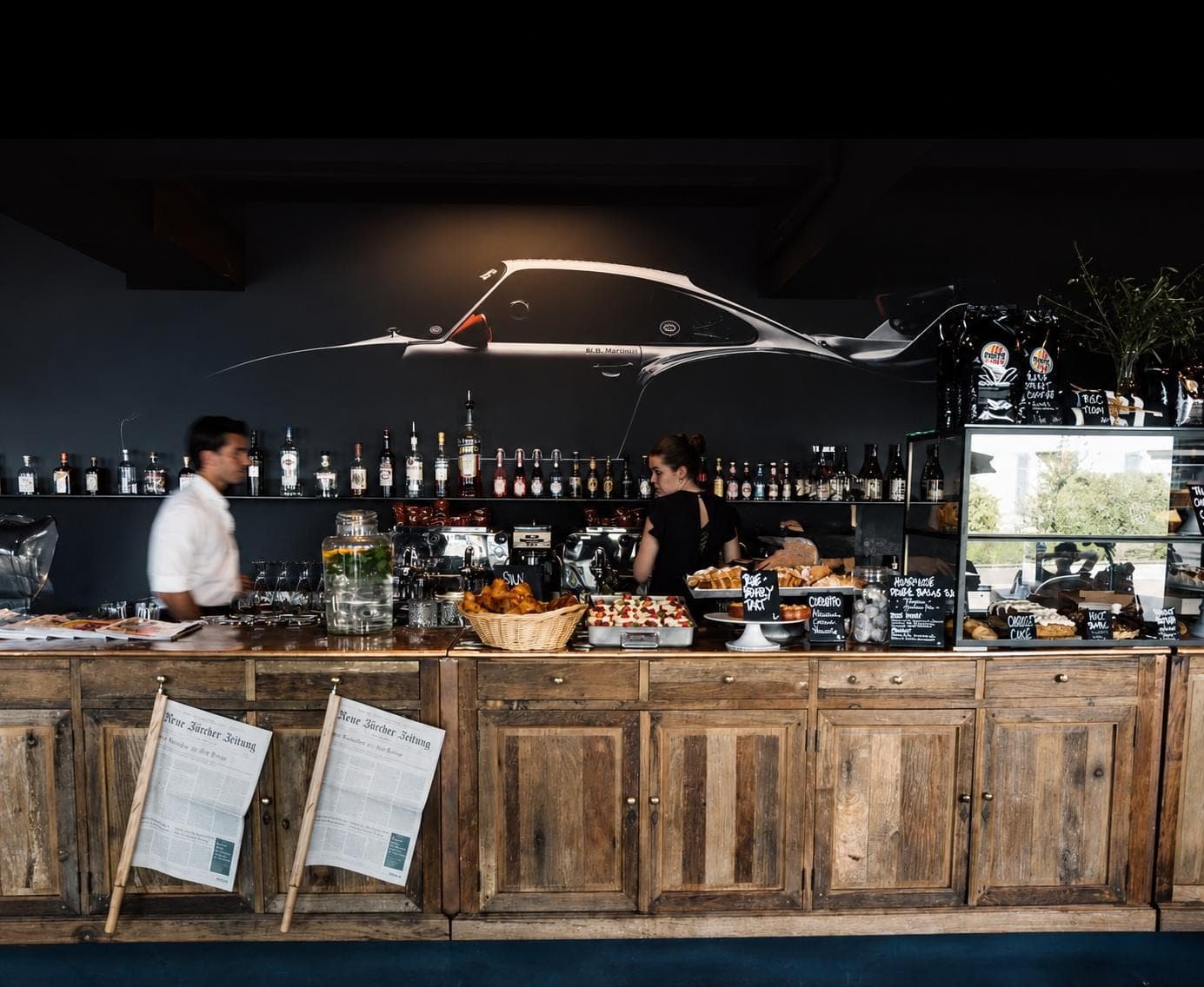 The bar at honeytree: a long reclaimed-wood counter set with pastries and coffee machines, a shelf of bottles behind it, and the silhouette of a classic sports car painted across the dark wall above.